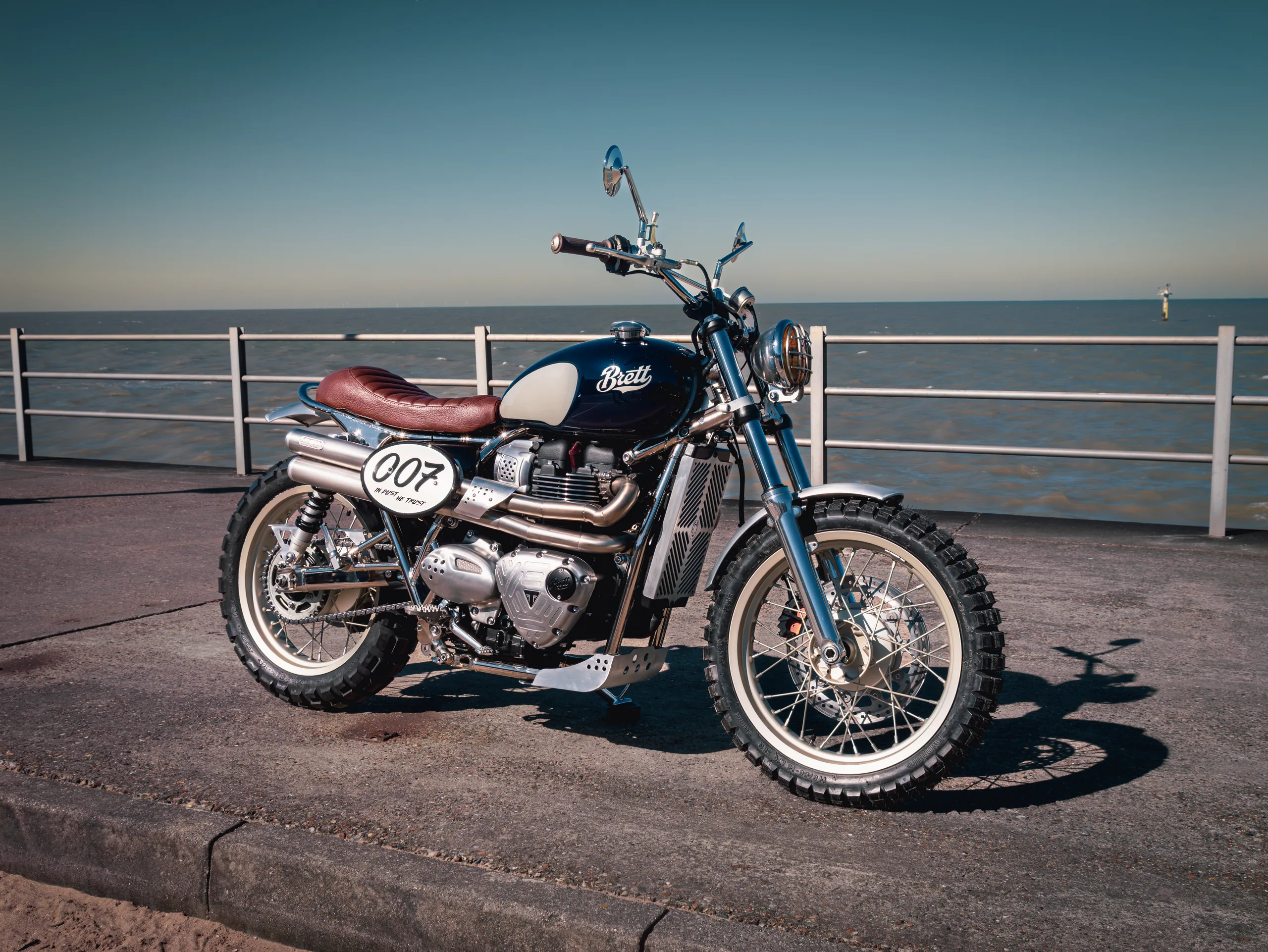 Triumph Legend FCR Original noire avec plaque 007 Brett Eyewear garée sur une promenade face à la mer