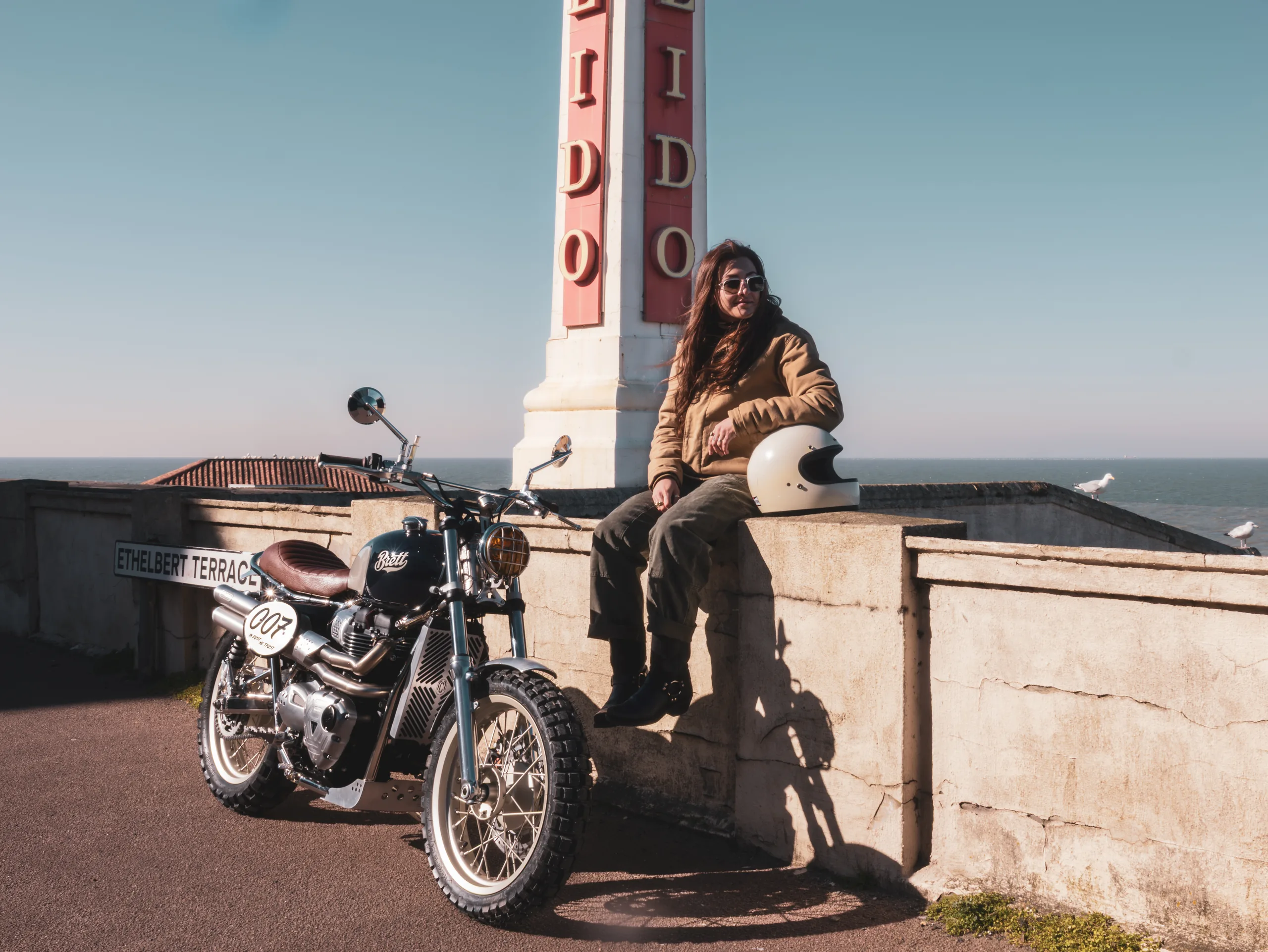 Motarde assise sur un muret près d'une tour Lido à côté d'une Triumph Legend FCR Original