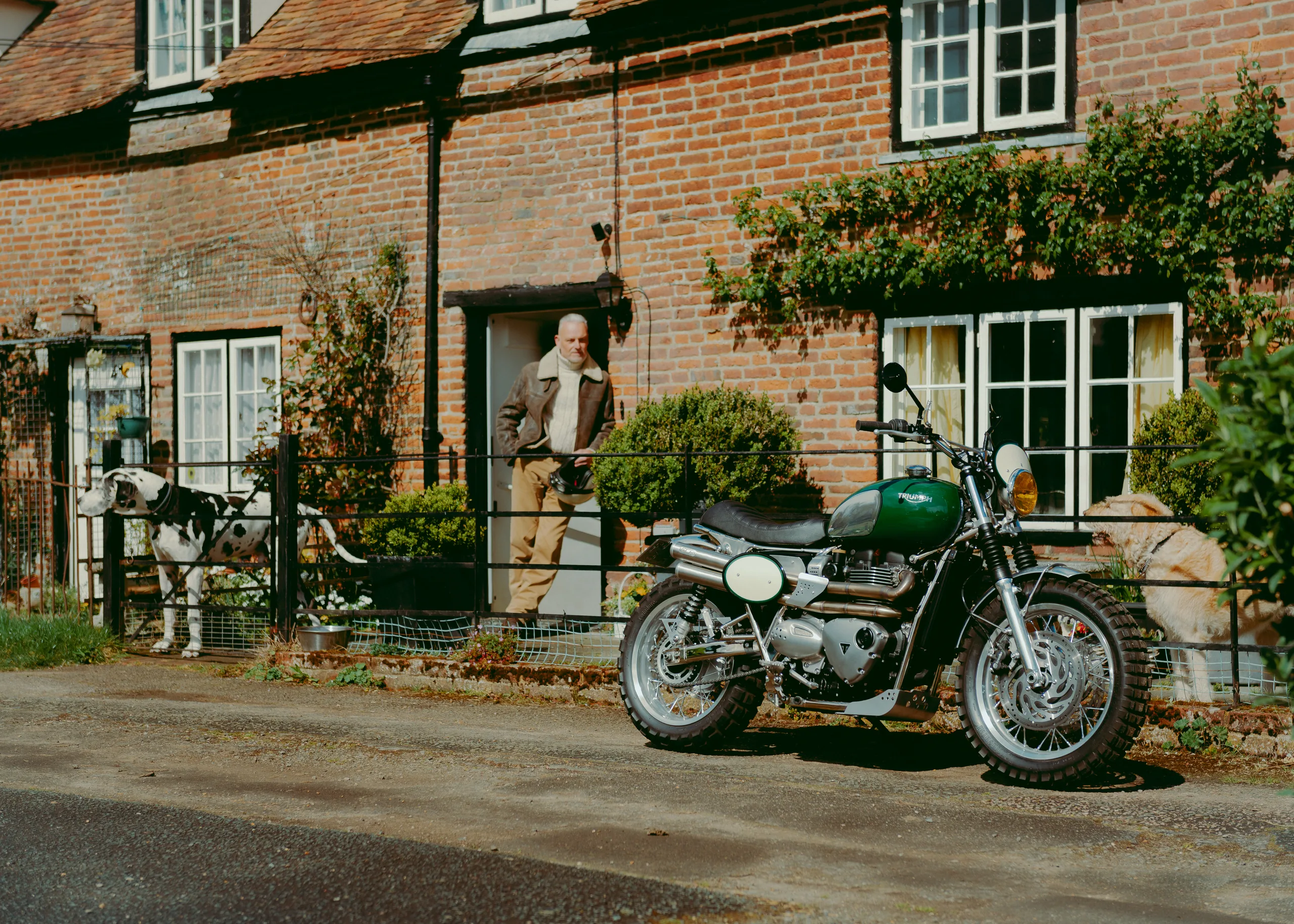 Triumph Legend FCR Original verte garée devant une maison anglaise en briques, un homme et un chien au portail