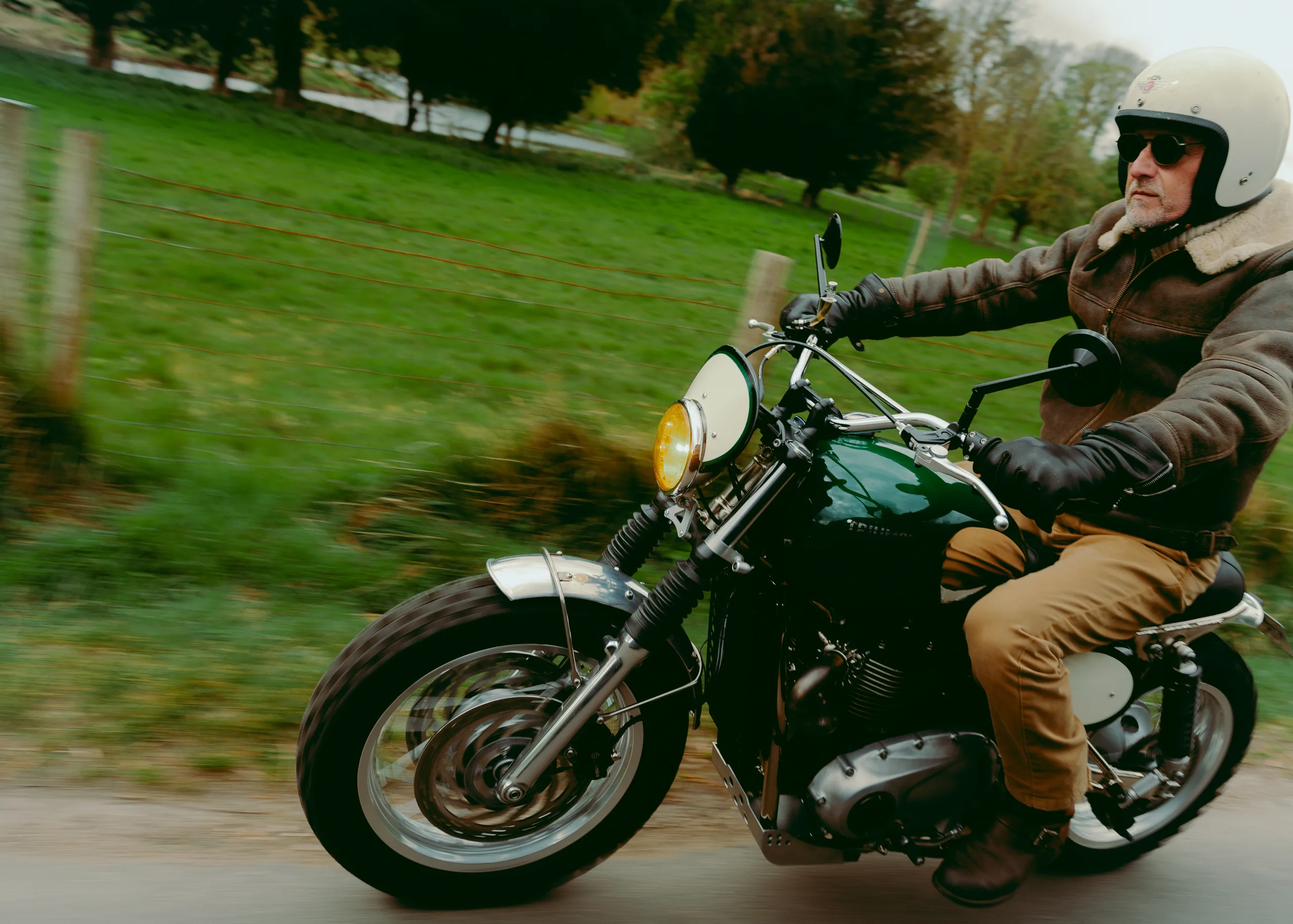 Motard en blouson roulant en Triumph Legend FCR Original verte le long d'un pré