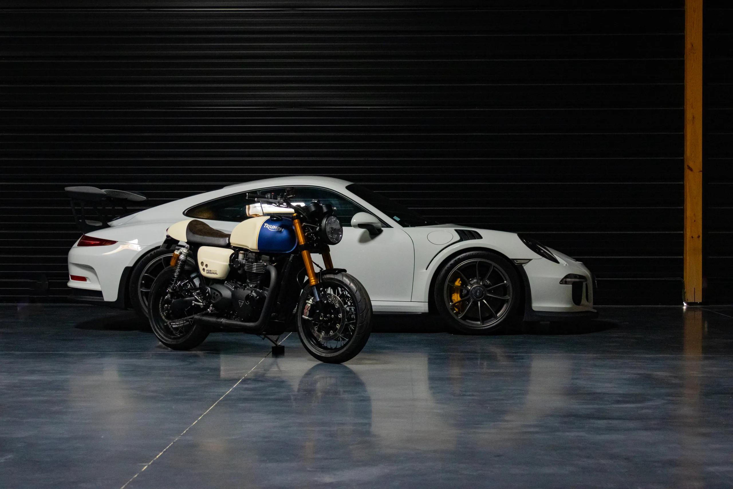 Porsche 911 blanche à aileron et Triumph Roadster Classic FCR Original dans un garage aux lames sombres
