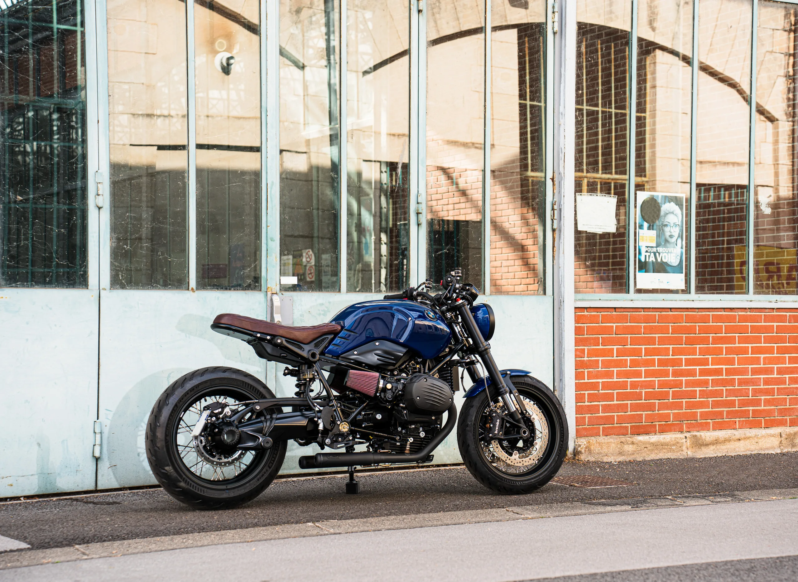 BMW R nineT Urban FCR Original bleue avec selle marron devant une façade d'atelier vitrée