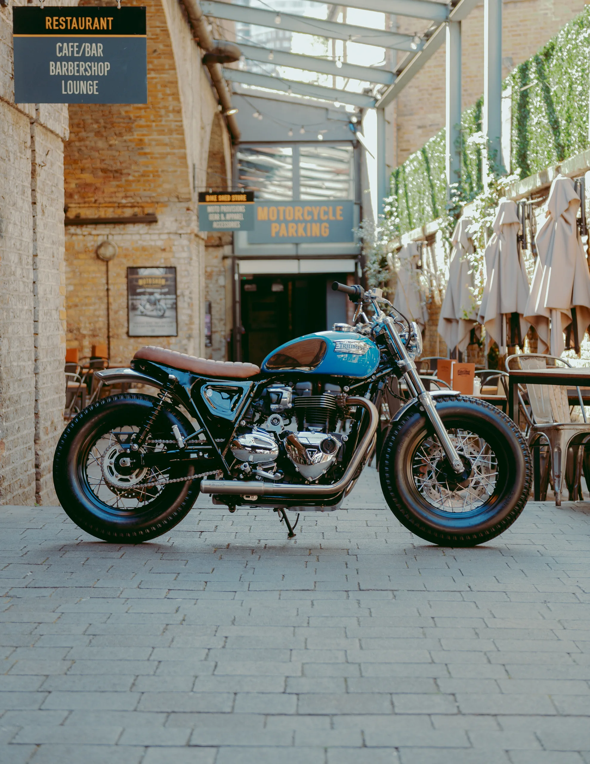 Triumph Fat Bike FCR Original turquoise avec selle marron dans une cour pavée près d'un parking moto