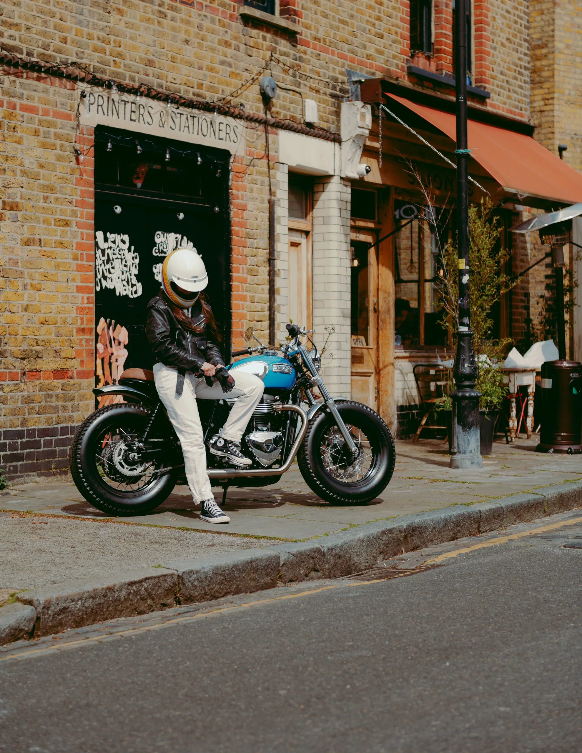 Motard au casque blanc sur une Triumph Fat Bike FCR Original turquoise devant une boutique londonienne
