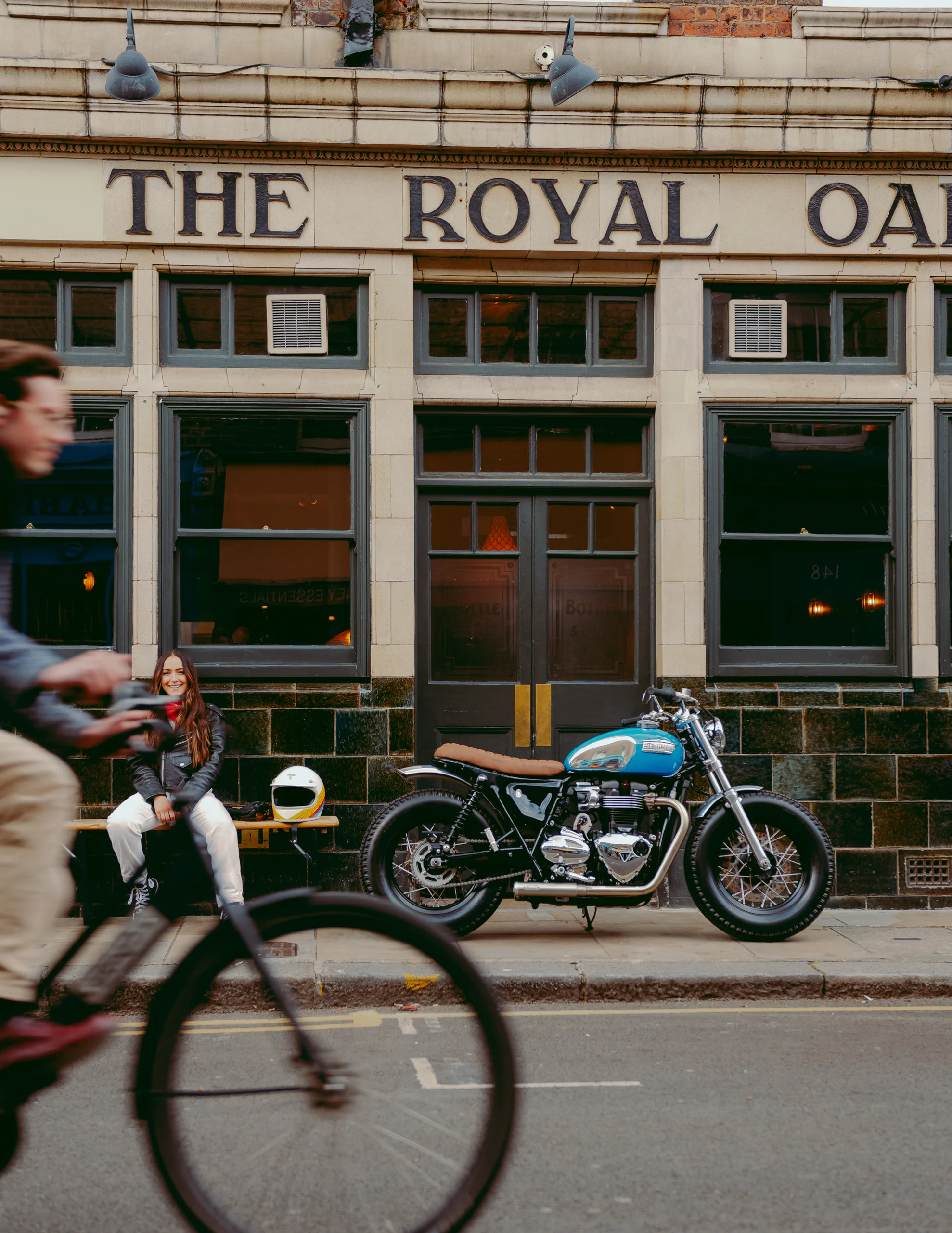 Triumph Fat Bike FCR Original turquoise devant un pub anglais, une motarde assise en terrasse