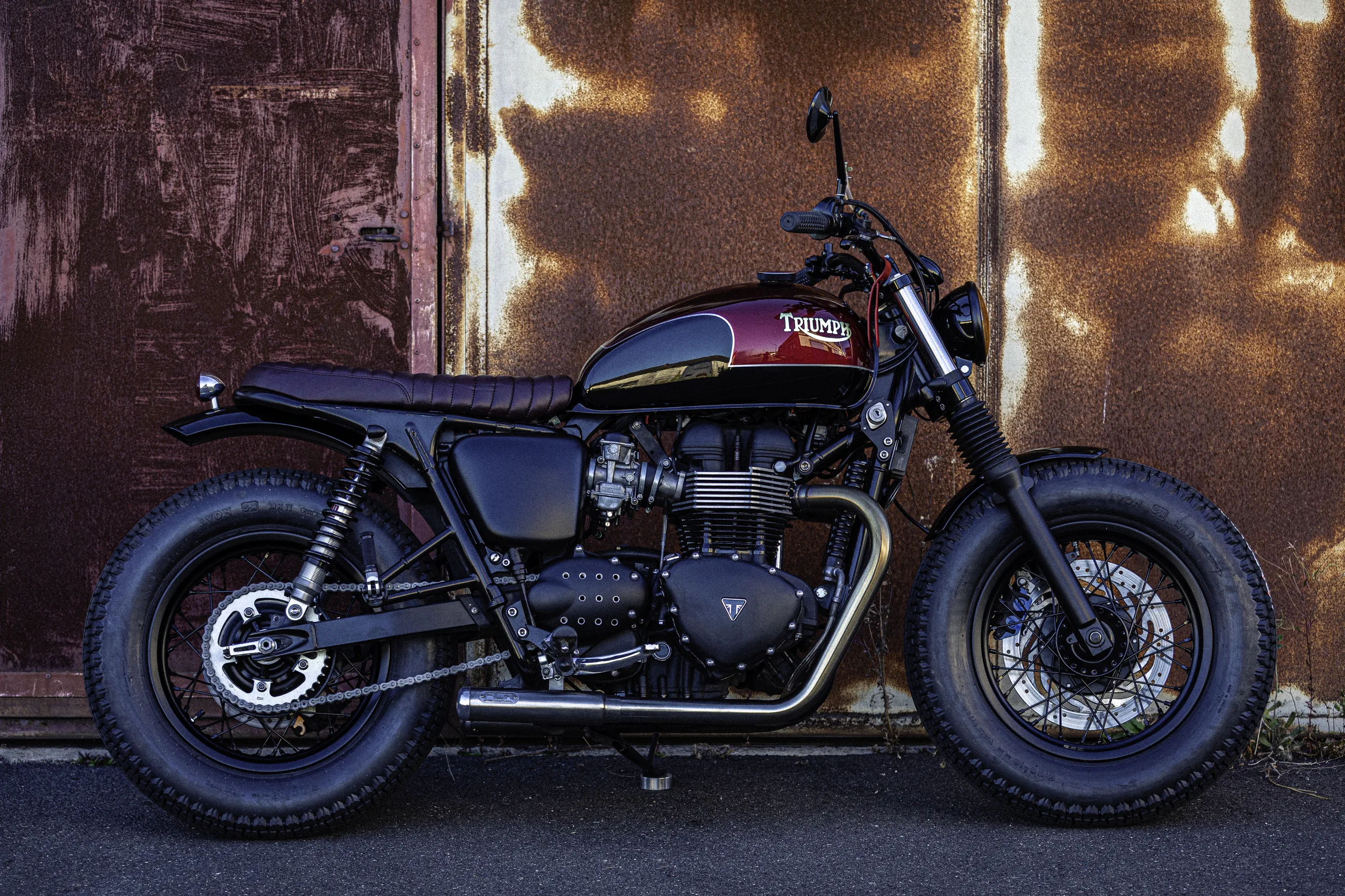 Triumph Fat Bike FCR Original bordeaux et noire garée devant un mur en métal rouillé