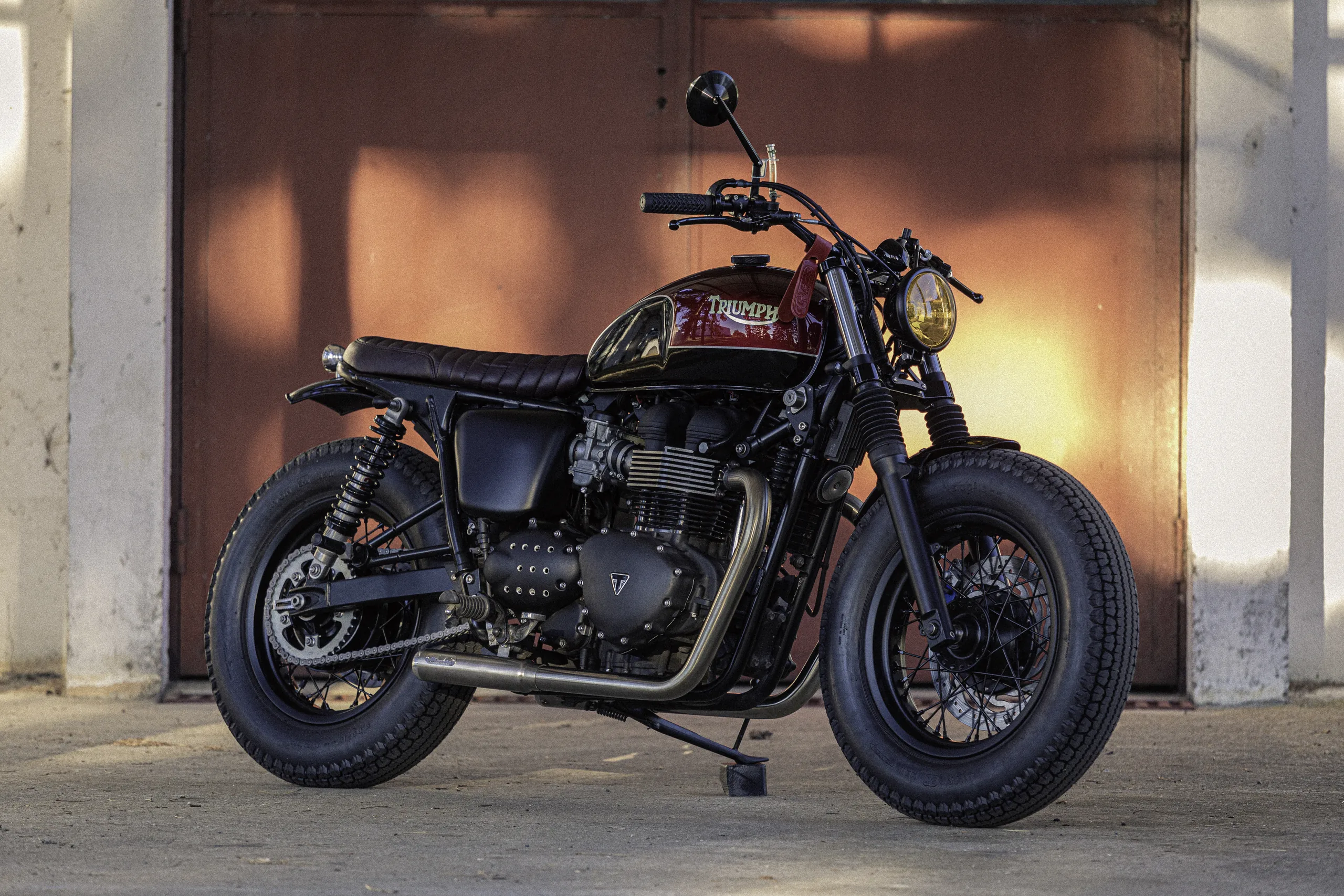 Triumph Fat Bike FCR Original bordeaux avec phare jaune devant une porte de garage au soleil couchant