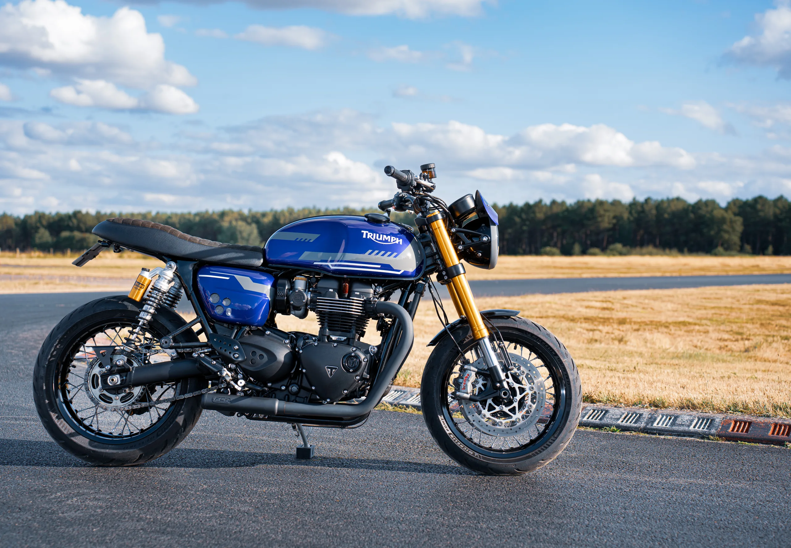 Triumph Thruxton RS Roadster Classic FCR Original bleue avec fourche dorée sur une route en campagne