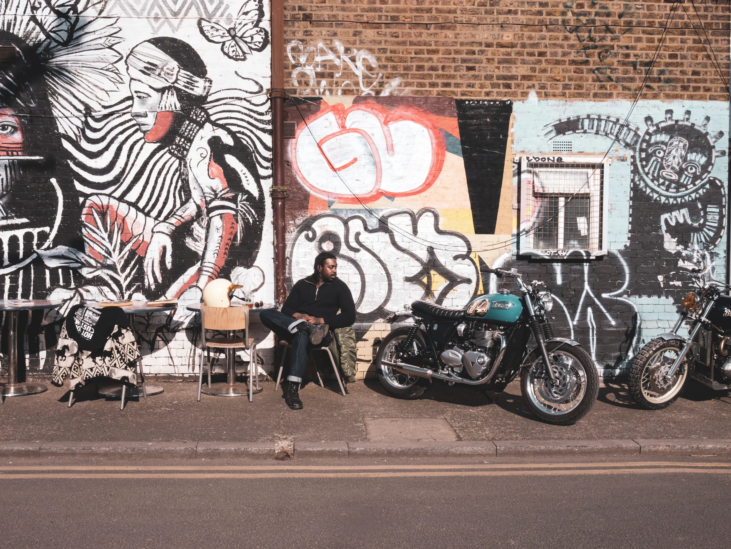 Homme assis en terrasse devant un mur de graffitis à côté d'une Triumph Ride & Chrome FCR Original turquoise