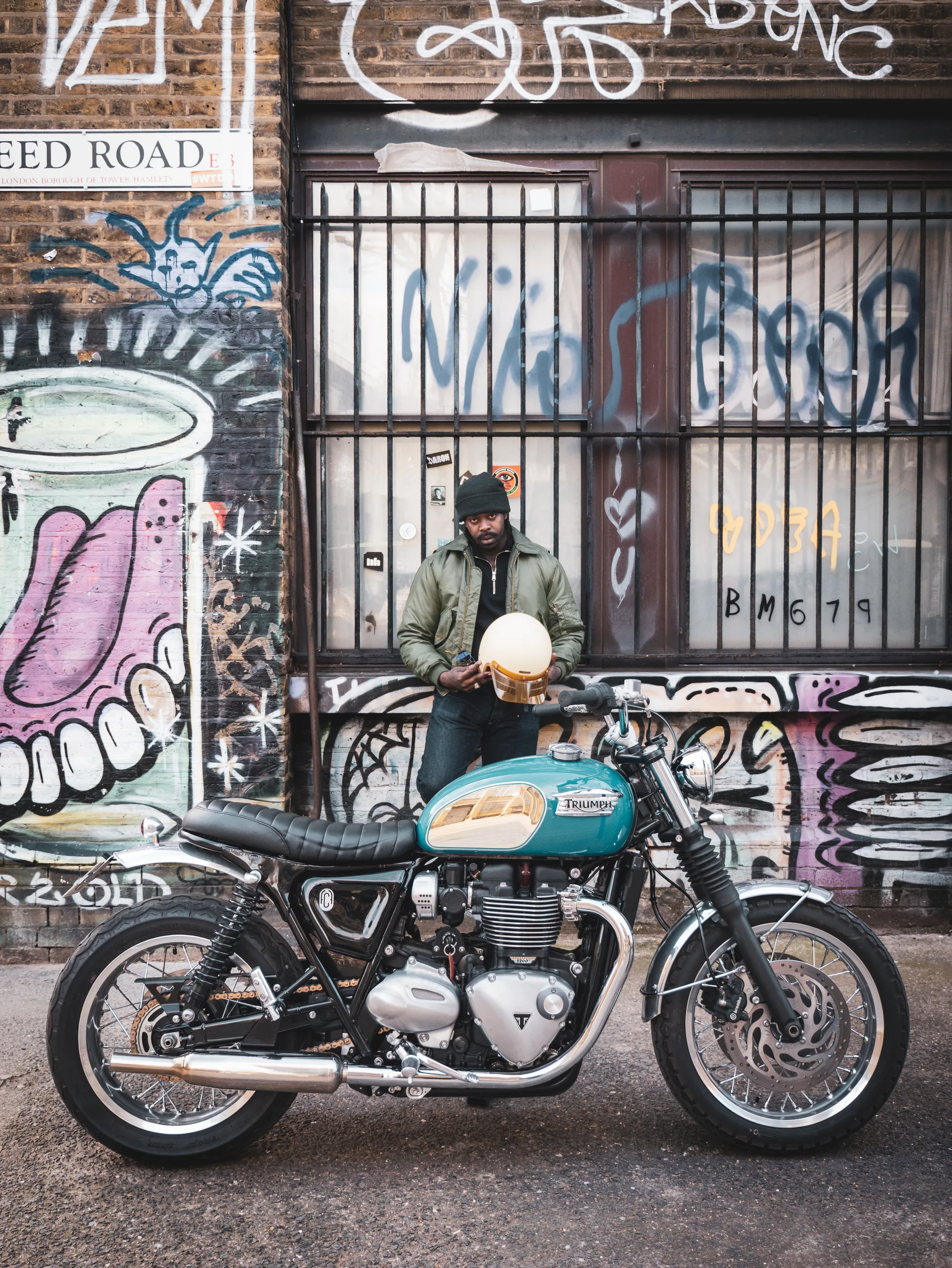 Motard avec casque devant un mur tagué à côté d'une Triumph Ride & Chrome FCR Original bleu turquoise