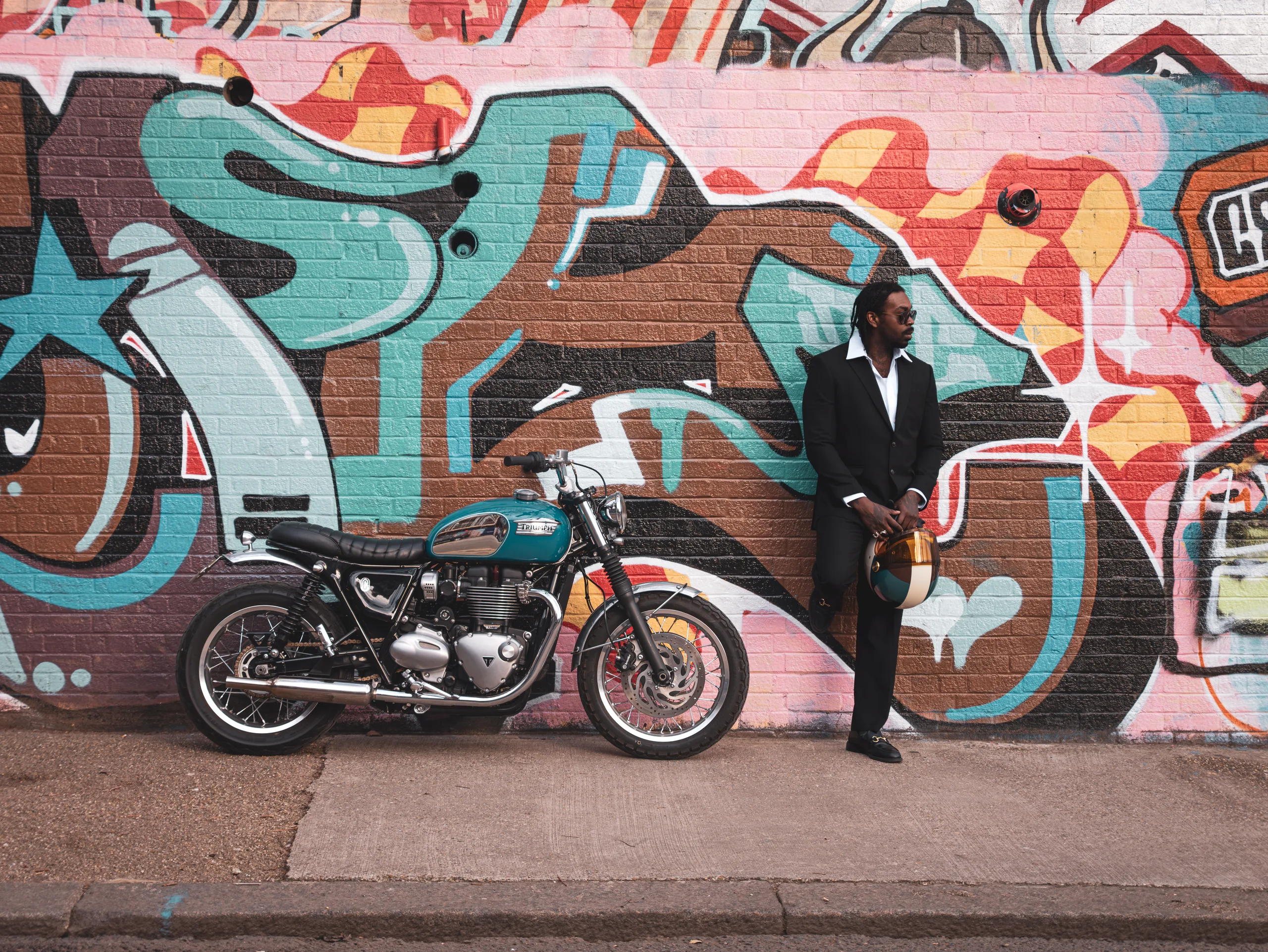 Homme en costume adossé à un mur de graffitis à côté d'une Triumph Ride & Chrome FCR Original turquoise