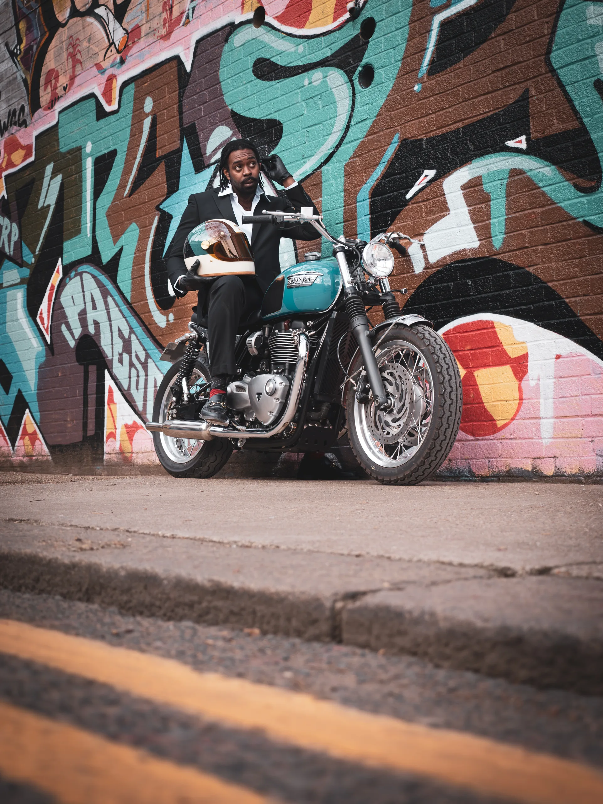 Motard assis sur une Triumph Ride & Chrome FCR Original turquoise devant un graffiti coloré