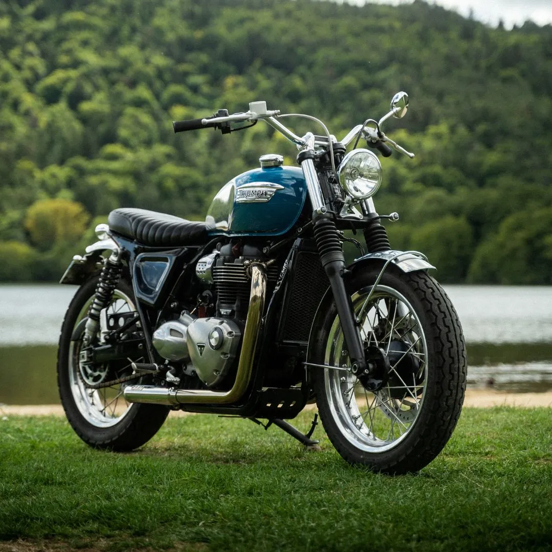 Découvrez la Triumph Speed Twin bleue vintage, une moto rétro devant un beau lac. Avec son design élégant et ses accents chromés, elle séduit les amateurs de motos classiques et de paysages naturels.