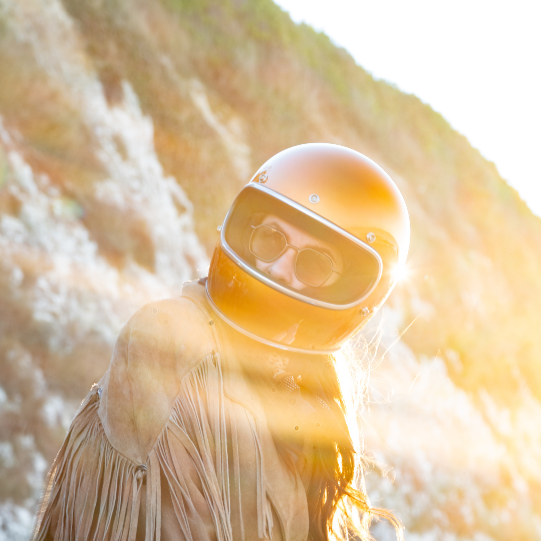 Portrait en contre-jour d’une pilote portant un casque doré et des lunettes Brett, ambiance vintage dans la lumière du soir.
