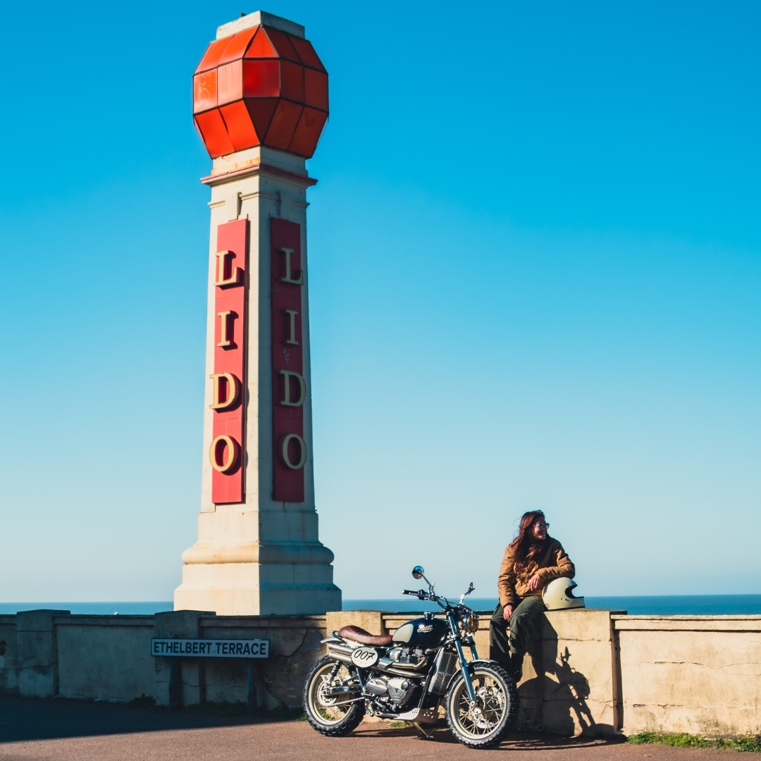 Triumph Legend FCR Original garée face à la mer, devant la colonne rétro "LIDO" de Margate, aux côtés de la pilote.
