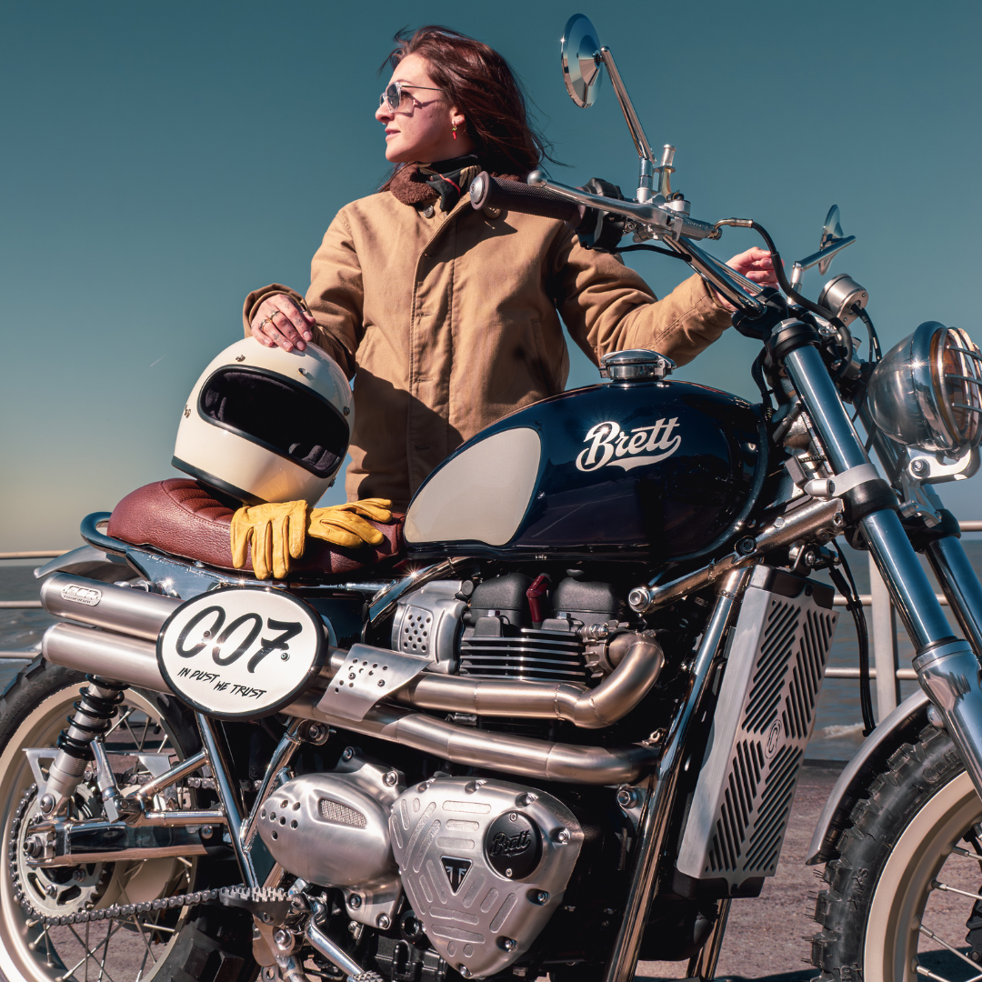 Portrait d’une pilote posant avec la Triumph Legend FCR Original, casque et gants posés sur la selle, devant le réservoir bleu Brett marqué "007".