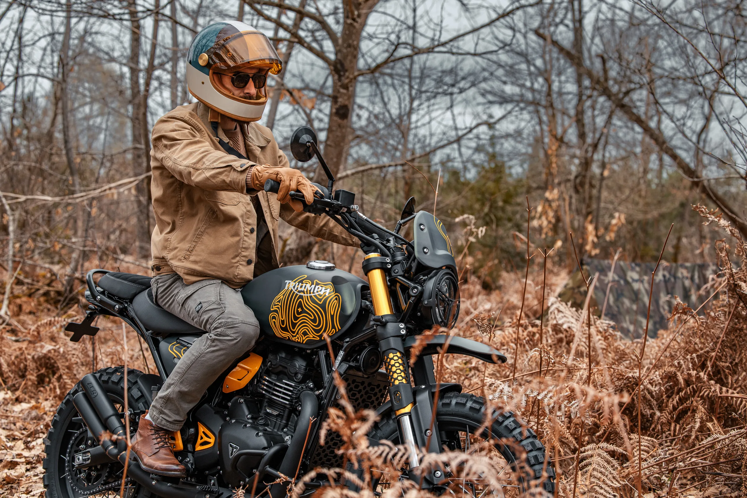 Pilote au casque vintage et veste camel sur une Triumph Scrambler 400 Escape FCR Original en forêt