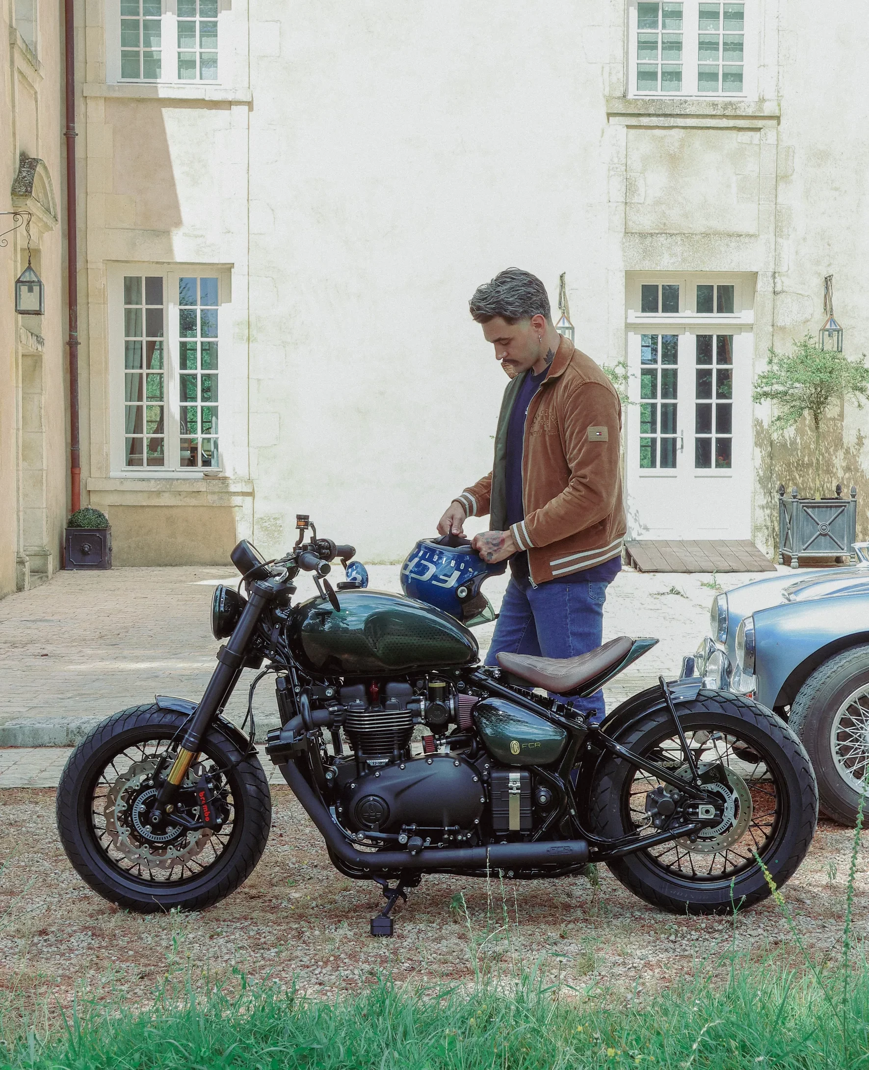 Pilote avec son Triumph Bobber préparé par FCR Original devant une maison en pierre
