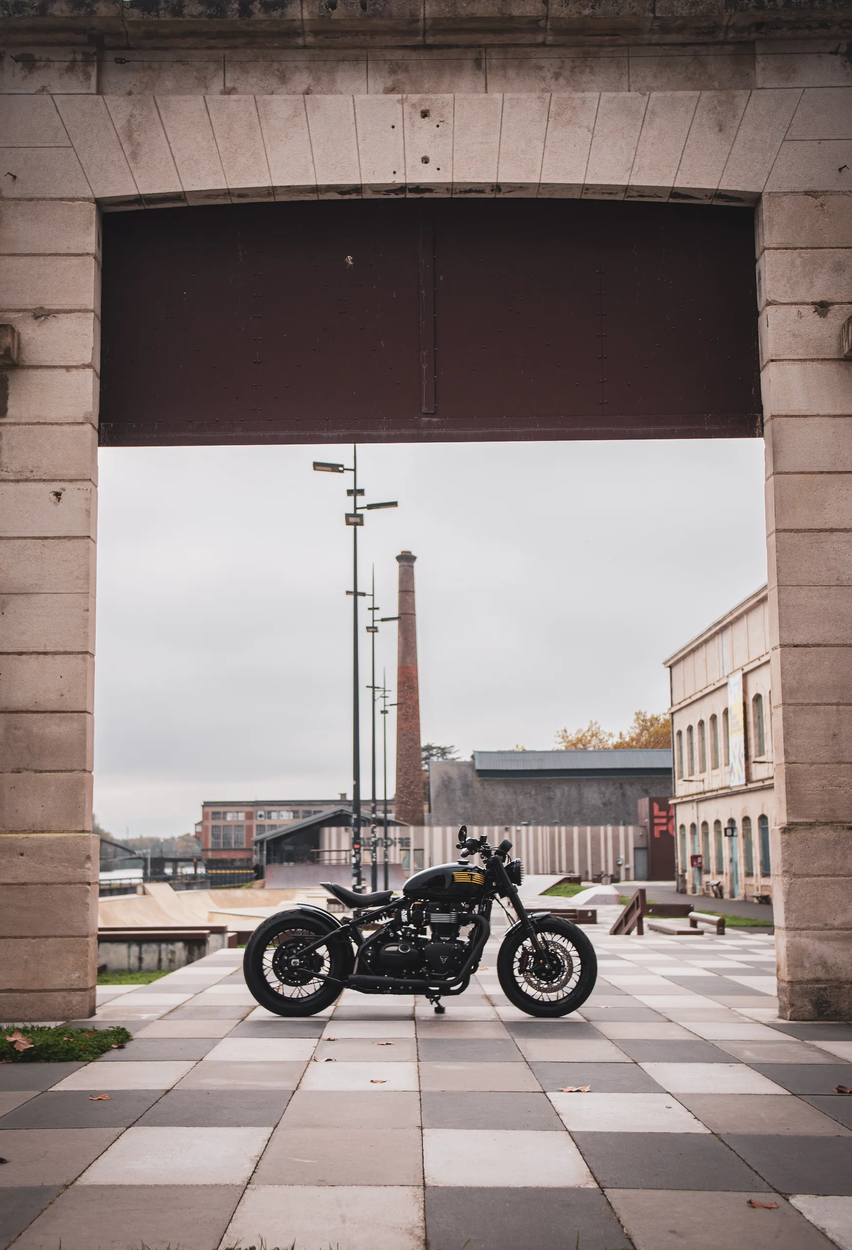 Triumph Bobber FCR Original encadrée par une arche en pierre, cheminée d'usine en arrière-plan